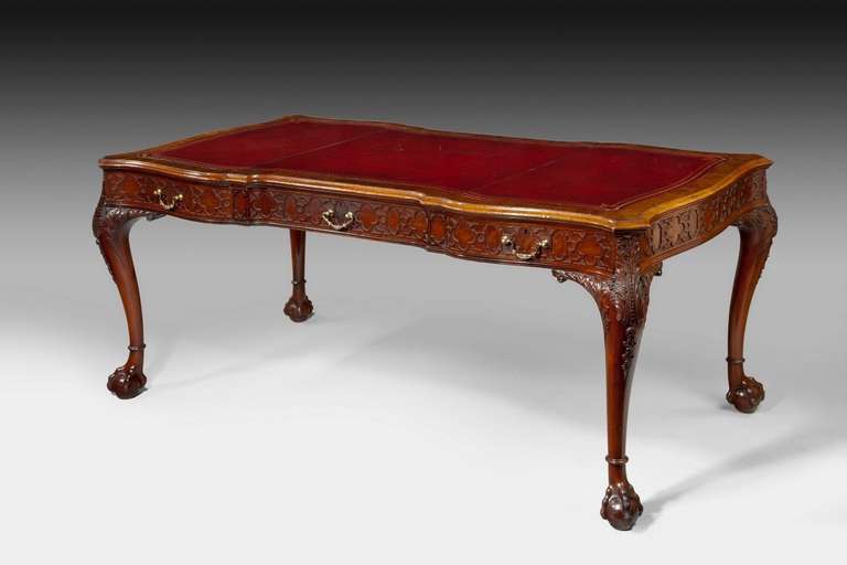 A Victorian mahogany library table, circa 1880, by Wright and Mansfield in George III manner, the rectangular red tooled leather inset top of arc-en arbelette form, above three drawers, with dummies to the reverse, all with blind fret carving, on
