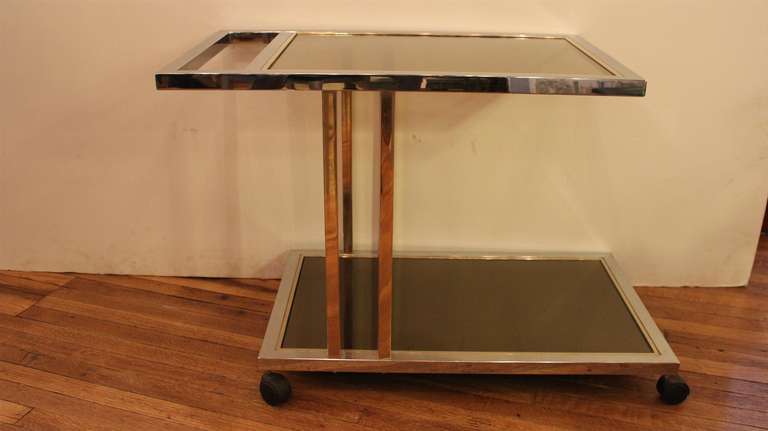 Mid-Century Modern chrome and smoky glass, asymmetrical two tier bar cart. Glass outlined in brass trim. This can be seen at our 302 Bowery locations in Manhattan.