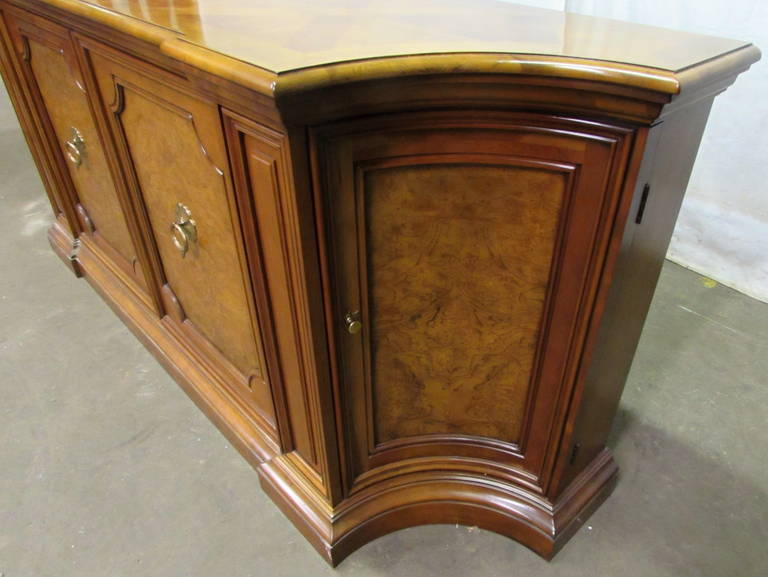 This is a large wooden sideboard with two-tone wood stain. There are two doors that have round brass pulls with floral embellishments. Each corner has a curved dark wood embellishment and the back two legs are set out farther than the front two.
