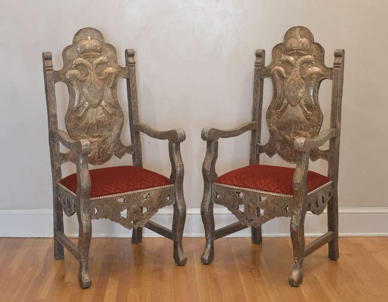 This pair of tall Chippendale style armchairs feature high, crested backs, undulating arms with scrolled hand rests, and seats embroidered in red velvet. Elaborate silver repoussé covers the front of each chair and on the back of each chair is the