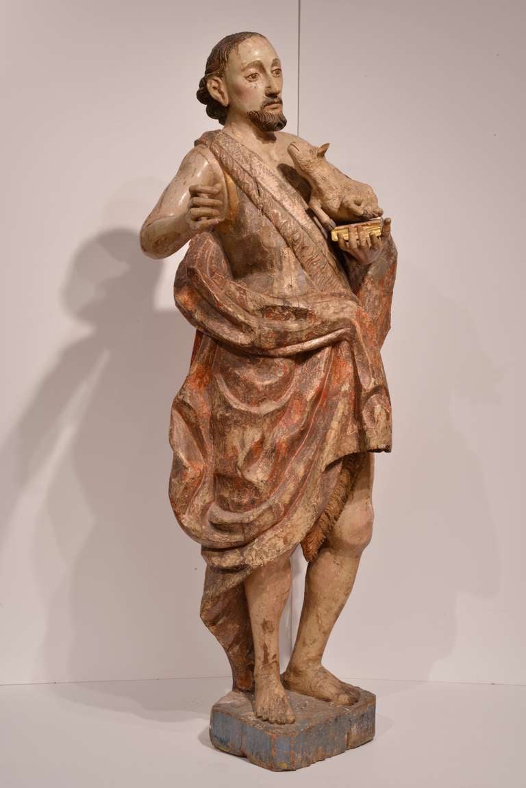 Dating to the late 17th/early 18th century, this wooden figure of St. John the Baptist is finely carved and embellished with striking detail. The figure has his right arm raised, and in his left arm he cradles a book and a lamb, whose little neck is