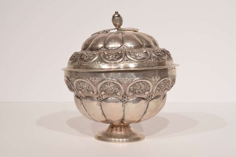 This set is comprised of the body and a lid. The body of this covered silver dish is low and circular, and each half of its surface covered with emphatic gadroons, crowned with semi-circular flourishes. The piece features floral detailing, with