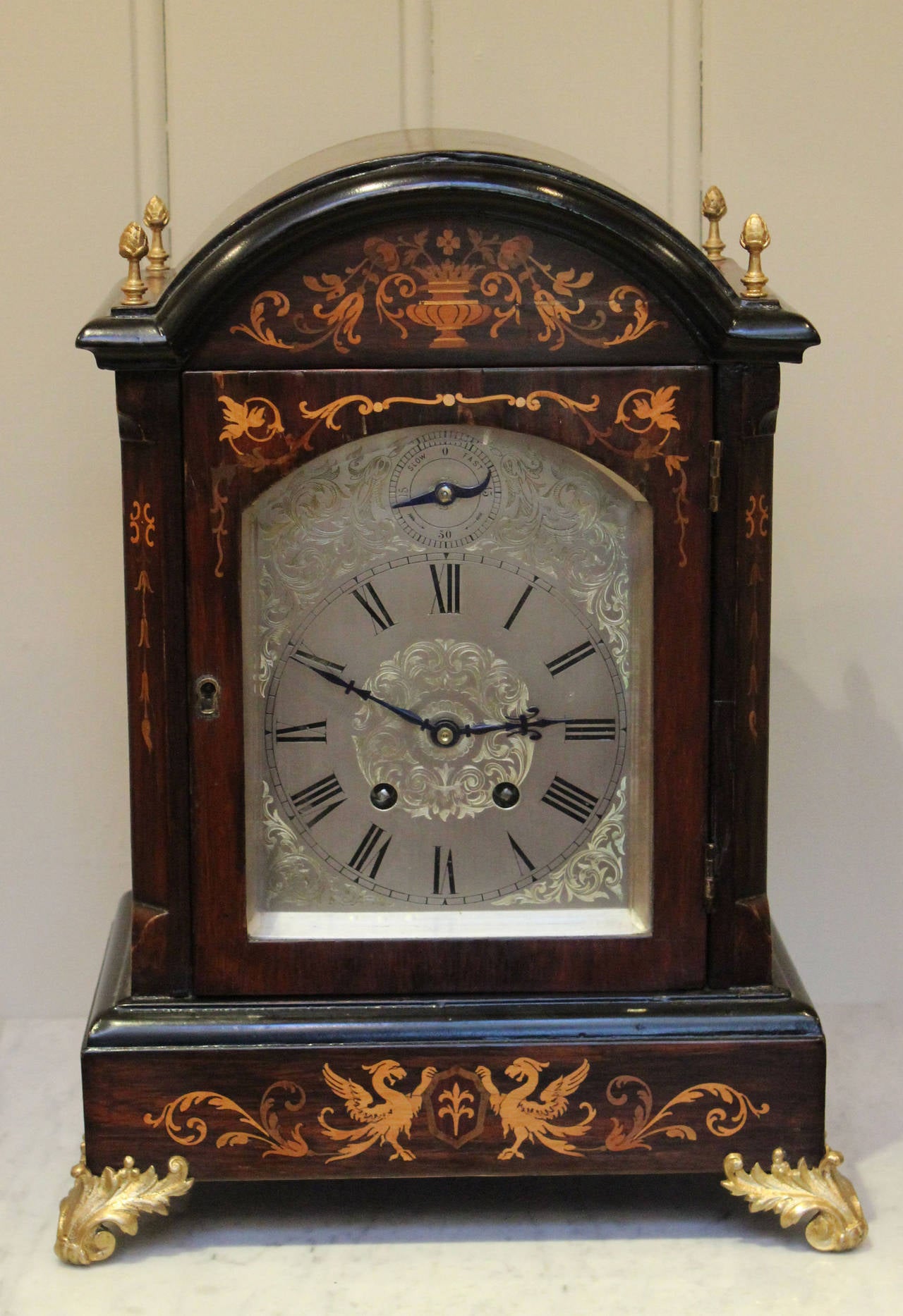 A substantial Rosewood Bracket Clock. It has an arch top case with marquetry inlay, brass corner finials and embossed bracket feet and bevel edge glass side panels. It has a well engraved silvered dial with a rise and fall to regulate the 8 day gong