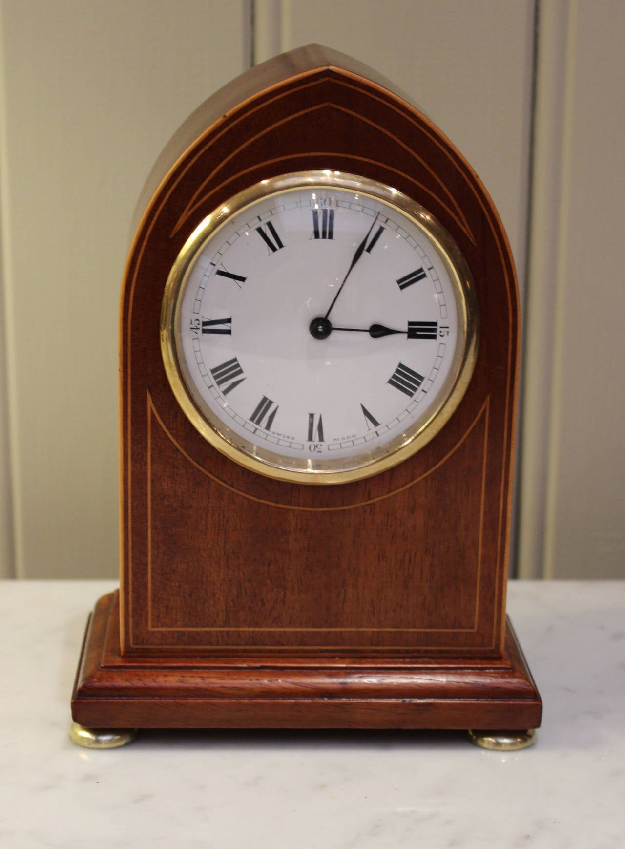 A small solid mahogany mantel clock, having a lancet shaped case inlaid with boxwood stringing and brass bun feet. It has a convex glass with an enamel dial and a Swiss 8 day lever platform movement.