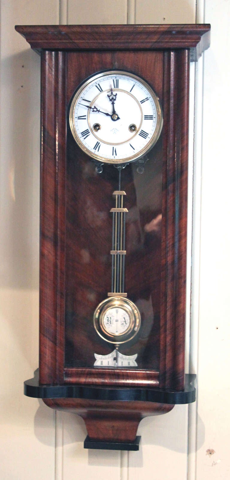 A slender wall clock of simple lines in a regulator style. It has a walnut case, with a shaped base and glass side panels and a detachable pediment and base finial.
The two piece enamel dial is marked by the maker Junghans, it has a gridiron