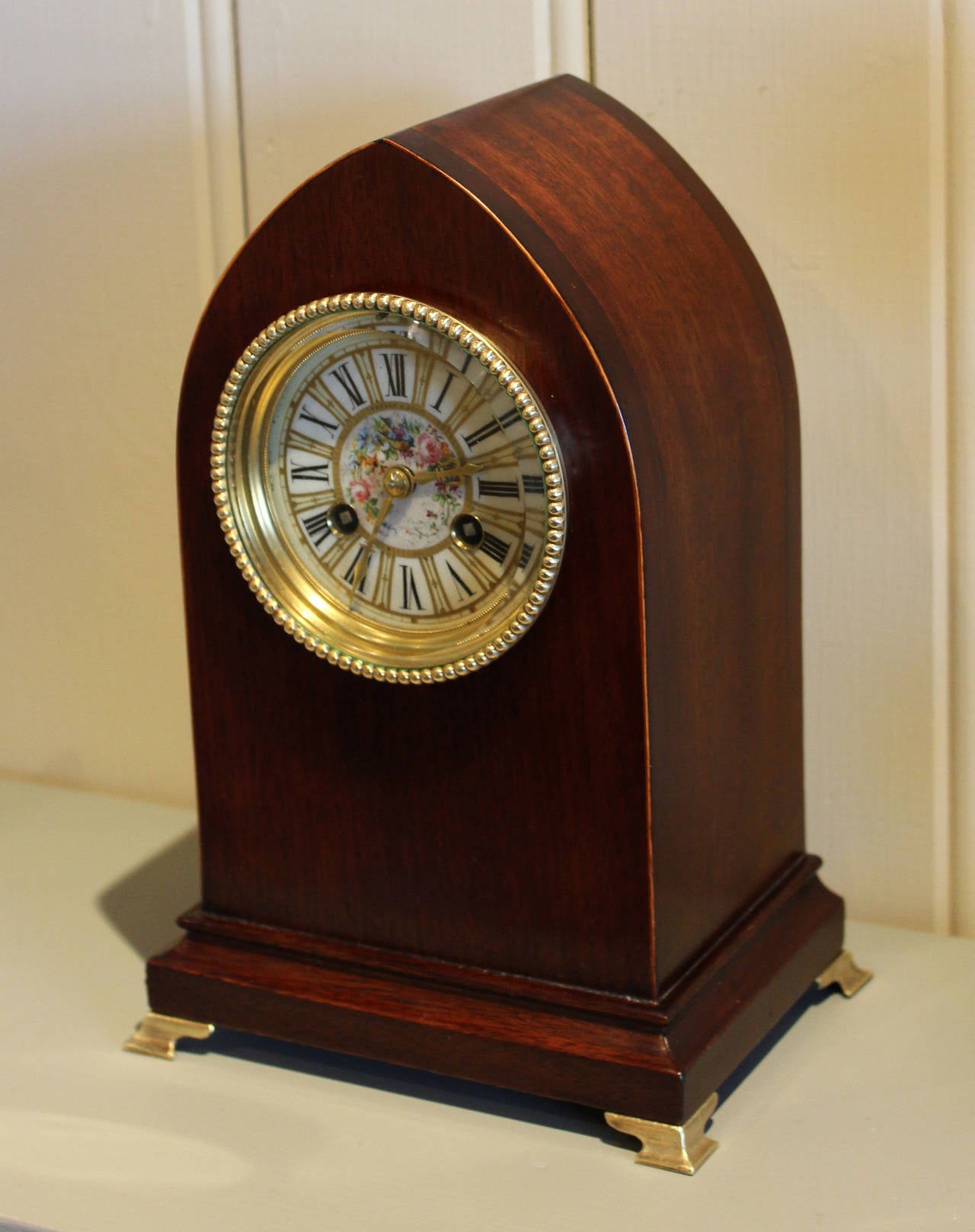 A solid mahogany mantel clock by Tiffany. The case has a lancet top with boxwood edging and brass ogee feet. It has a beaded brass bezel with a thick bevel edge glass and a ceramic painted enamel dial that is decorated with birds among floral