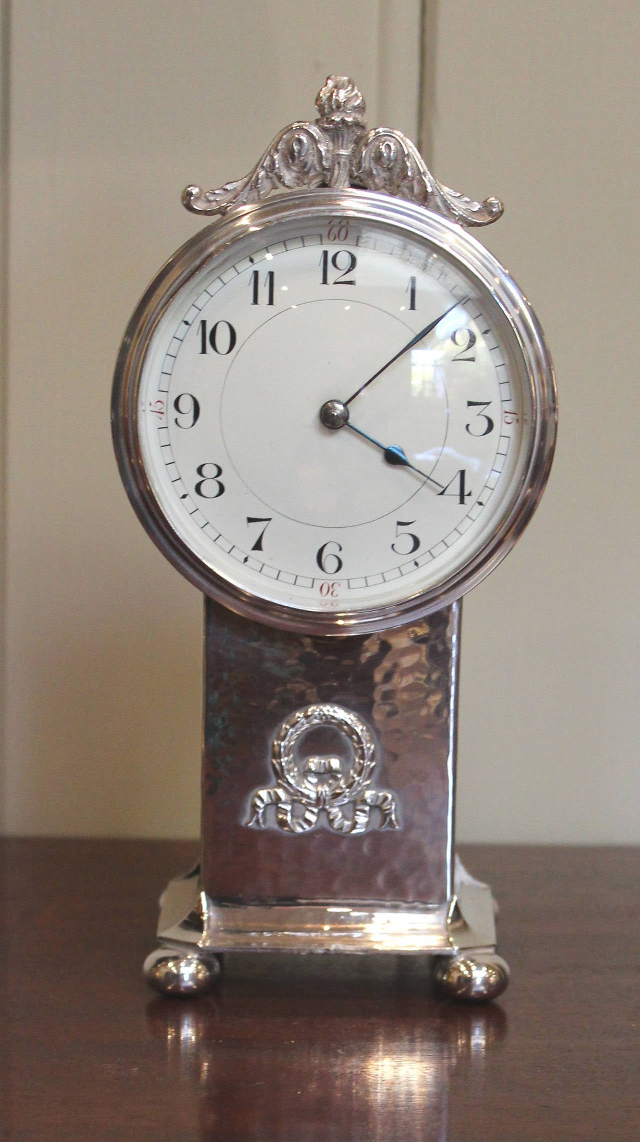 A silver plated mantel clock in the form of a drum mounted on a pedestal. It has a hammered case with a cresting mounted on a circular drum containing the movement. The pedestal has a swag ribbon and wreath and a molded base with bun feet. The