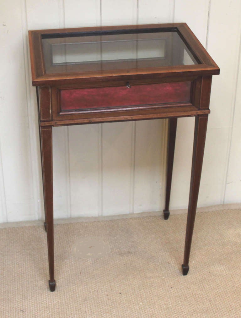 A solid mahogany bijouterie display table. It has a hinged top which is cross banded with satinwood and inlaid with boxwood stringing. Th sides are glazed with a red velvet lined base, standing on tapering supports.