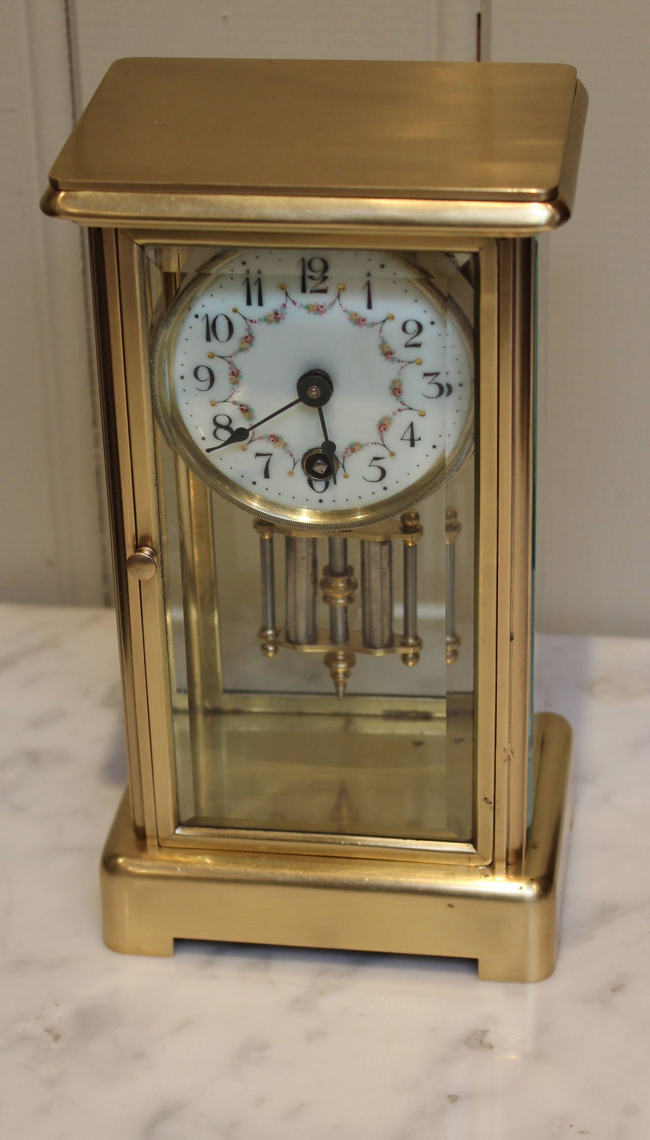 A small, timepiece four glass clock, having a plain brass case with bevel edge glass to the sides, and front. It has an 8 day pendulum movement, with a floral enamel and a simulated mercury pendulum.