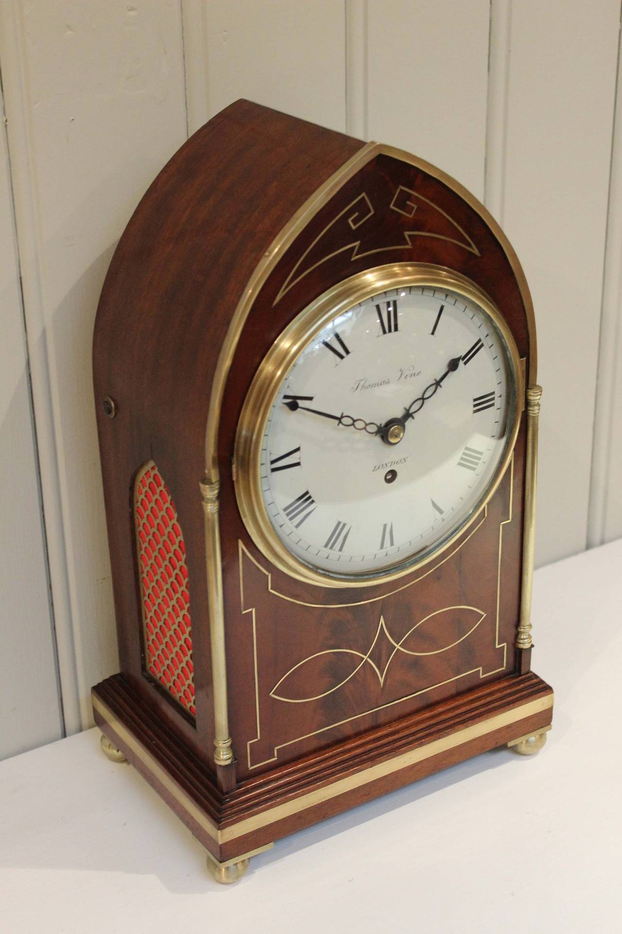 A fine mahogany timepiece bracket clock, dating to the Regency period. It has a lancet shape case with geometric brass inlay , turned brass columns and military style banding around a stepped base, with brass ball feet and arched brass fish scale