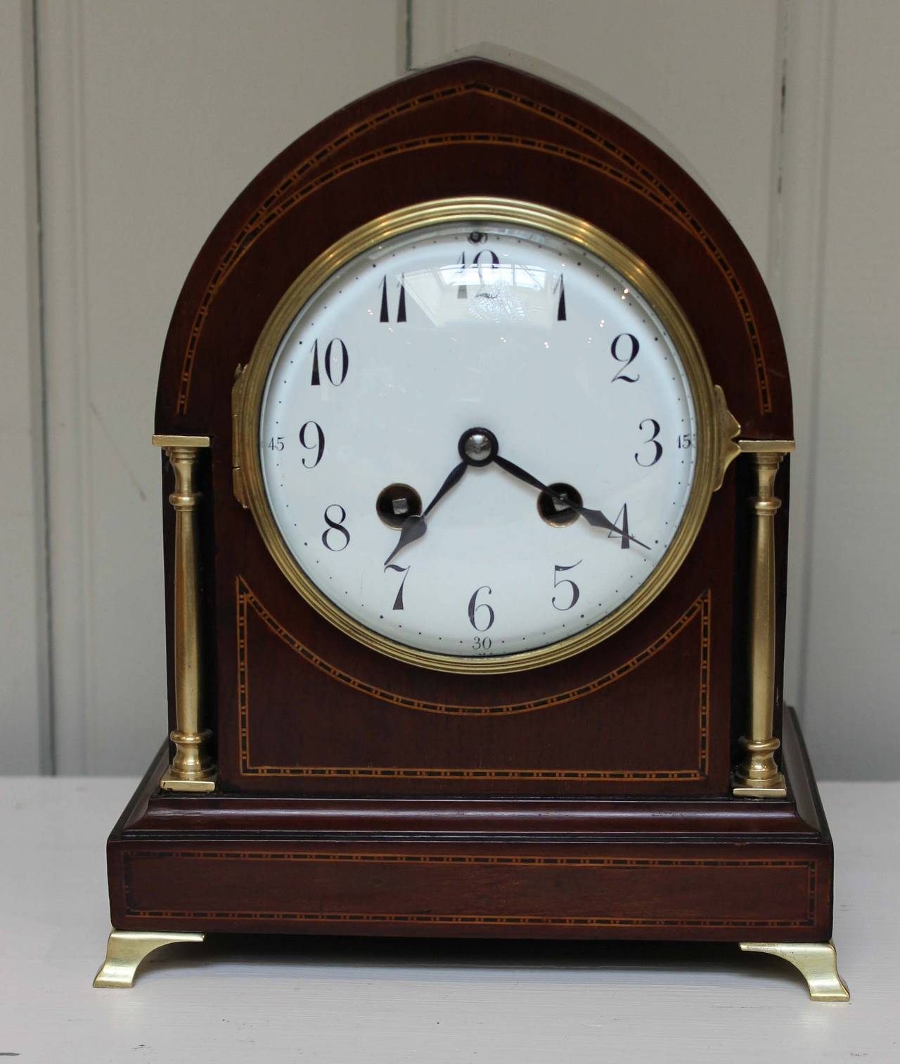 A solid mahogany mantel clock. It has a lancet top shaped case, boxwood stringing and brass side columns and brass bracket feet. The enamel dial has a convex bevel edge glass. It has a French eight-day pendulum hour and half hour gong striking
