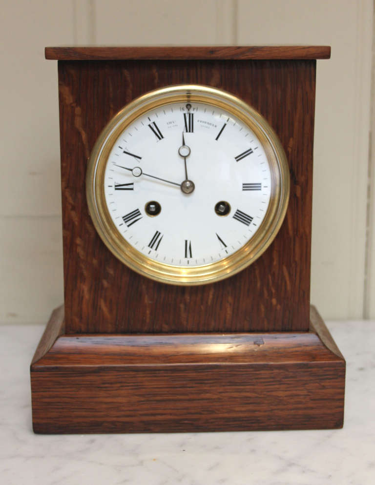 A small, solid oak mantel clock of simple rectangular form. It has a figured oak case, with a white enamel dial with moon hands. It is signed by the original suppliers, Charles Frodsham of London and Paris. It has an eight day French made movement