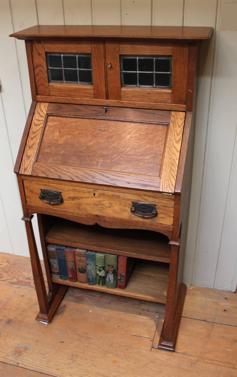 A small proportioned solid oak Arts and Crafts bureau. It has a top two door cabinet with leaded glass, a bureau section with a fitted interior and a bookcase base.
