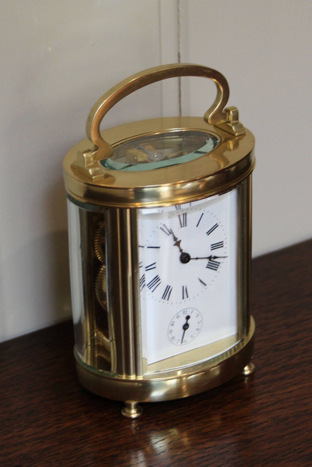 An oval brass carriage clock dating to the end of the 19th Century, which is larger than the standard timepiece models. It has four bevel shaped glasses and a bevel edge oval top glass. The enamel dial has a subsidiary alarm dial. The 8 day movement
