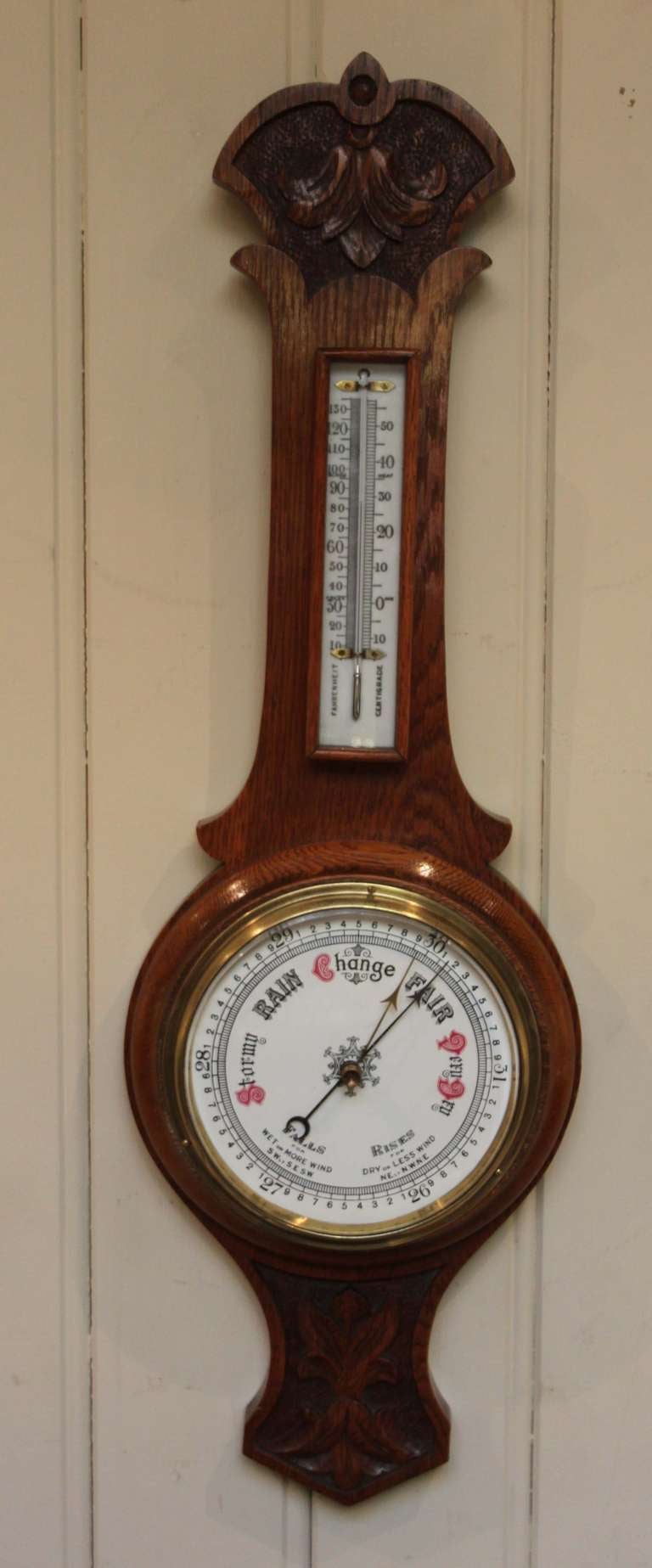 Solid golden oak aneroid banjo barometer having a carved top and bottom with a white dial dial and a mercury thermometer