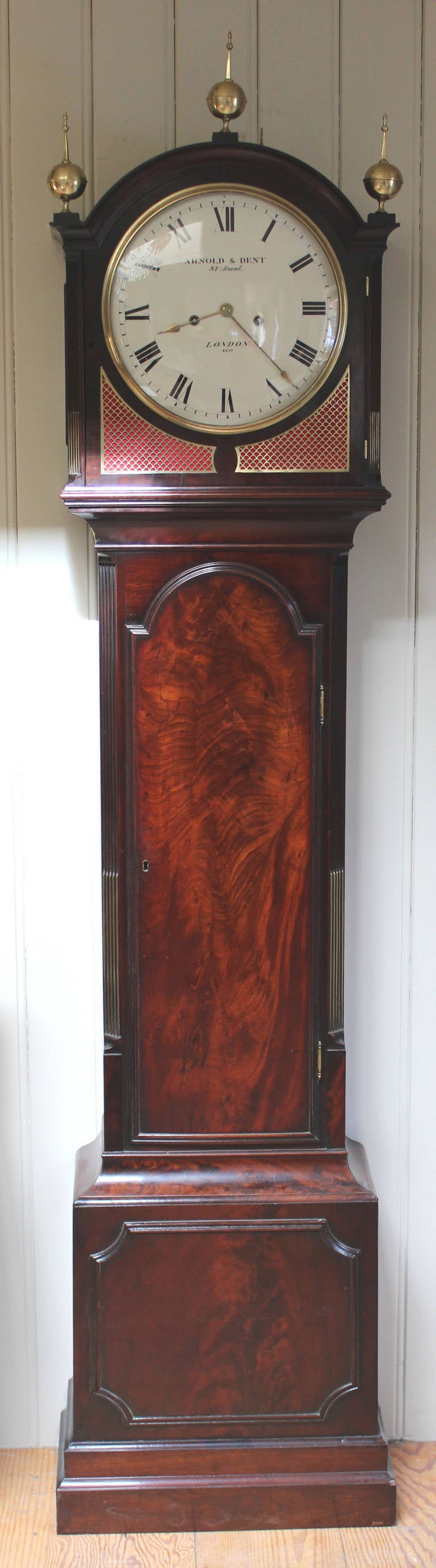 A fine and original, well proportioned mahogany longcase clock made by Arnold and Dent numbered 460. It has a break arch hood mounted by three brass finials, brass fish scale fret panels and a white convex dial with a convex glass. The case has a
