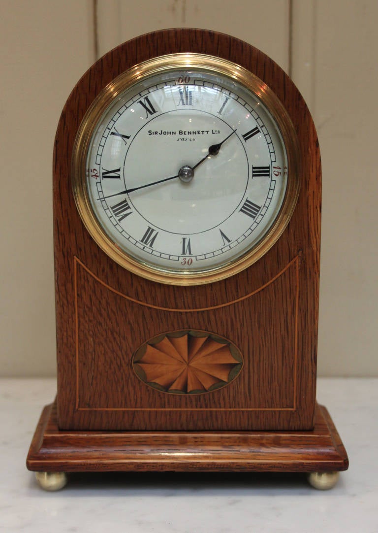 A small solid oak mantel clock, with boxwood stringing and satinwood inlays. The enamel dial is signed by the retailer Sir John Bennett Ltd. of London and Paris It has a French eight-day lever platform movement.