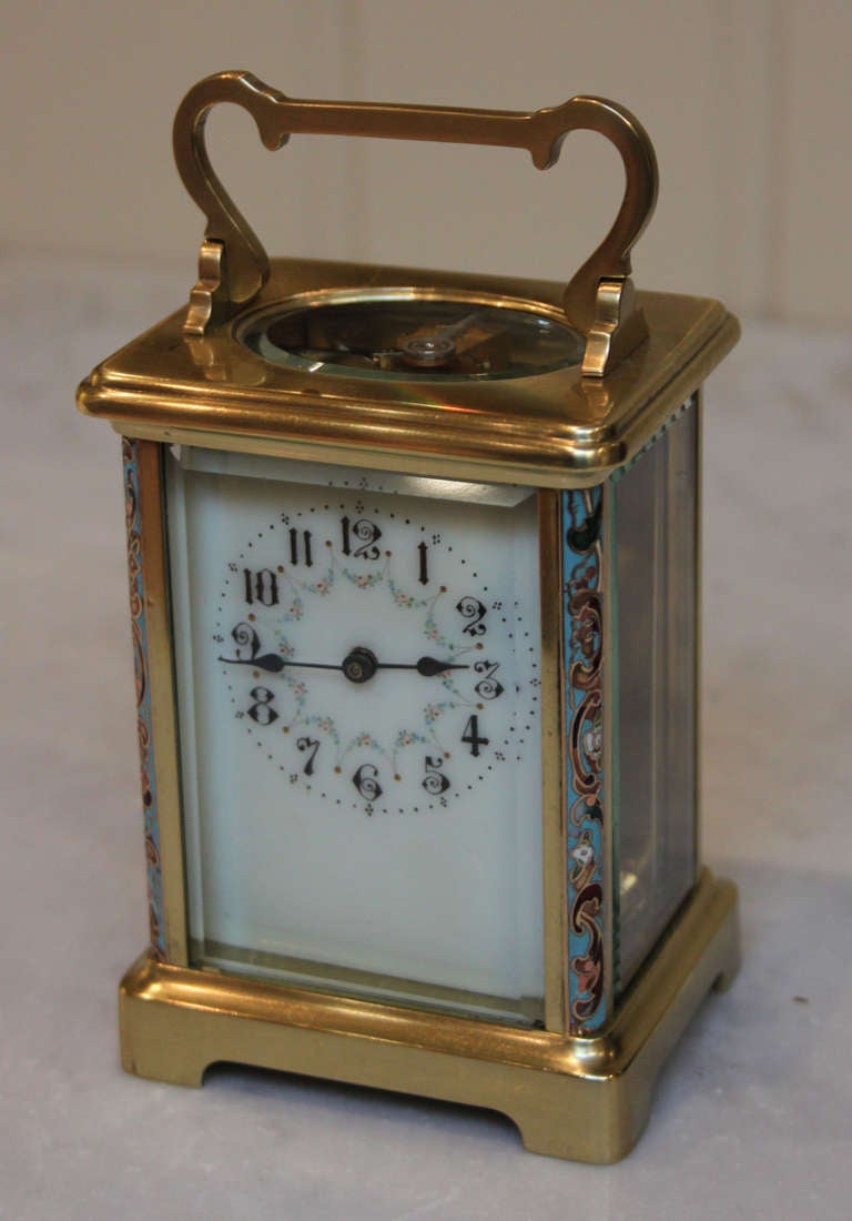 A Timepiece carriage clock in a brass case. It has a corniche case, with an oval top glass and champleve enamel decoration to the front corners. It has n ivory coloured enamel dial with floral decoration. The 8 day movement has a lever platform