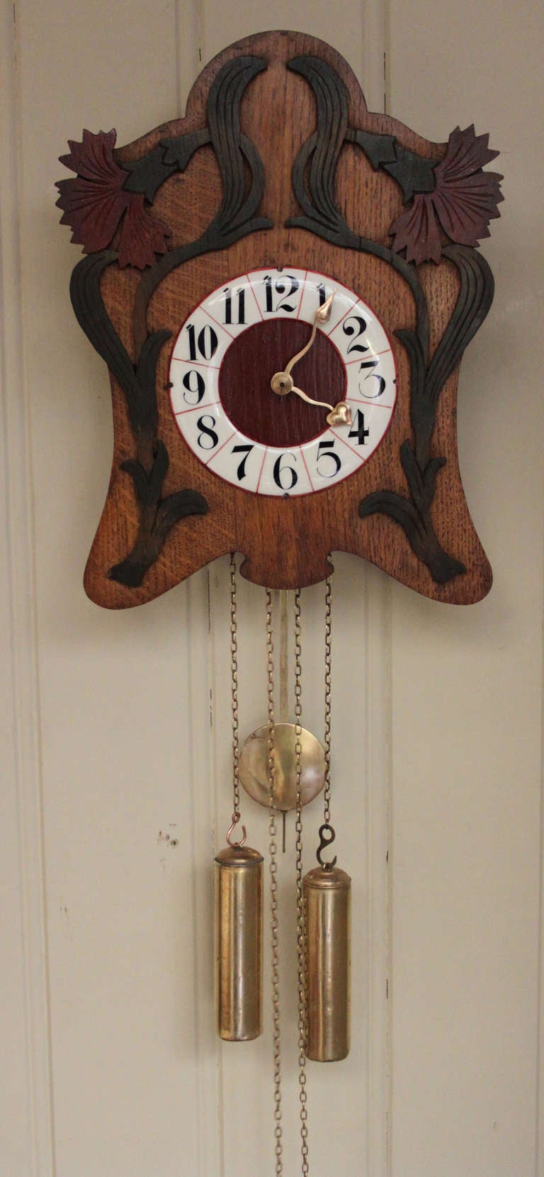 An unusual Art Nouveau wall clock from the Black Forest. It has an oak front with carved, colored applied vine leaves, and an enamel chapter ring with red lines and block numerals. It has a wooden plated movement with bass weights and pendulum that