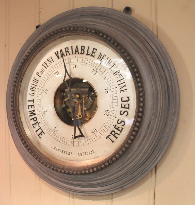 An unusually large aneroid barometer. It has a grey painted stepped turned frame, with a beaded bezel. It has an engraved silvered dial and a visible brass movement.