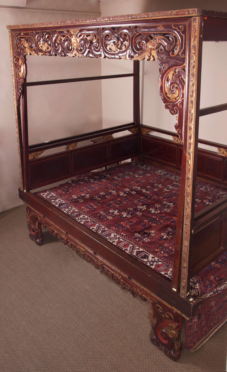 A sumptuous, four poster marriage bed, carved in teak and decorated with red lacquer and gilt. Incorporating a carved exterior canopy decorated to the front with a pair of large carved dragons' heads and with three bats to the centre, and an