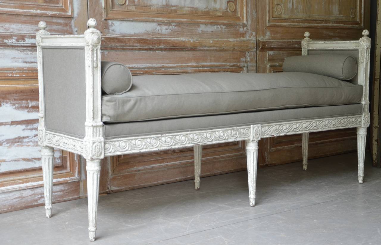 19th century narrow, long bench in Louis XVI style with armrest, richly carved side rails and upholstered seat with down cushion and bolster pillows in linen,
France, 1880.