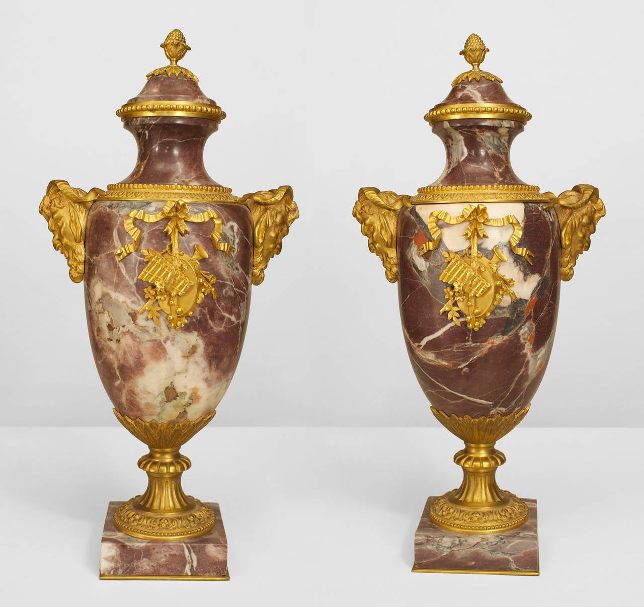 Pair of French Victorian Neoclassical style rosso levanto marble urns with gilt bronze ram head sides and acorn finial top with musical relief trim supported on a square base. (PRICED AS Pair)
