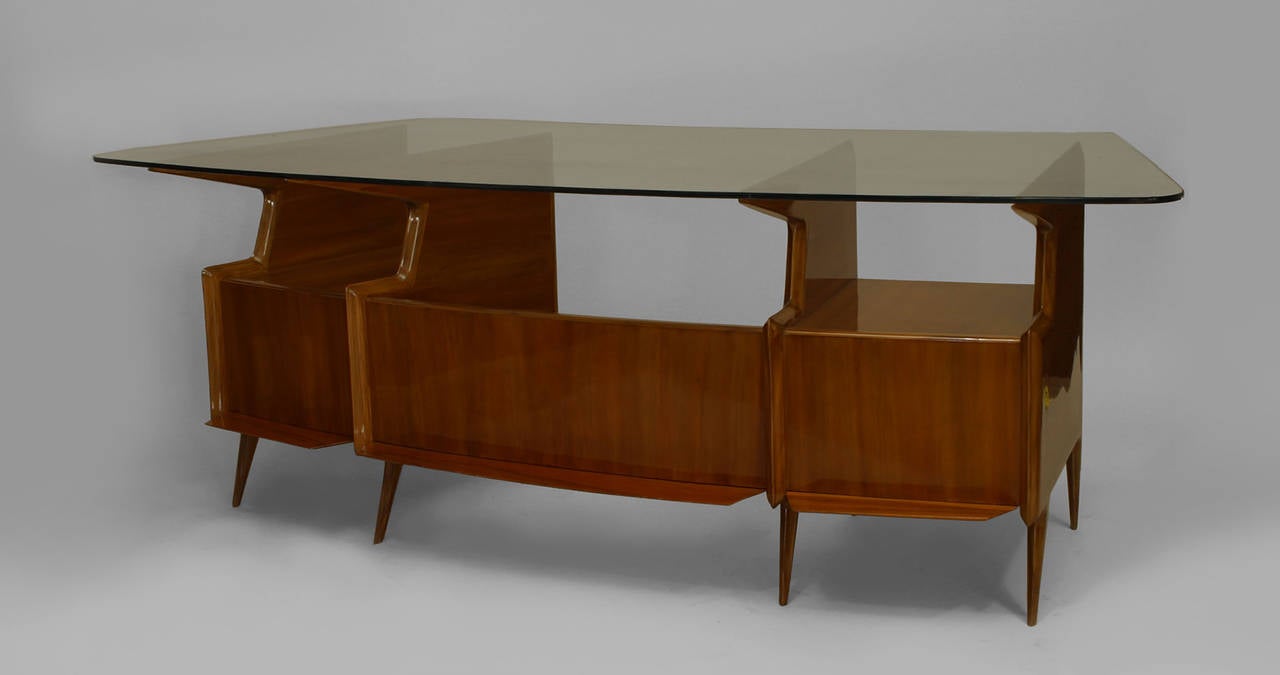 Designed by Italian Mid-Century Modern artist Gio Ponti, this mahogany, bowed desk features a glass top over open side shelves and two drawers separated with a vanity panel.