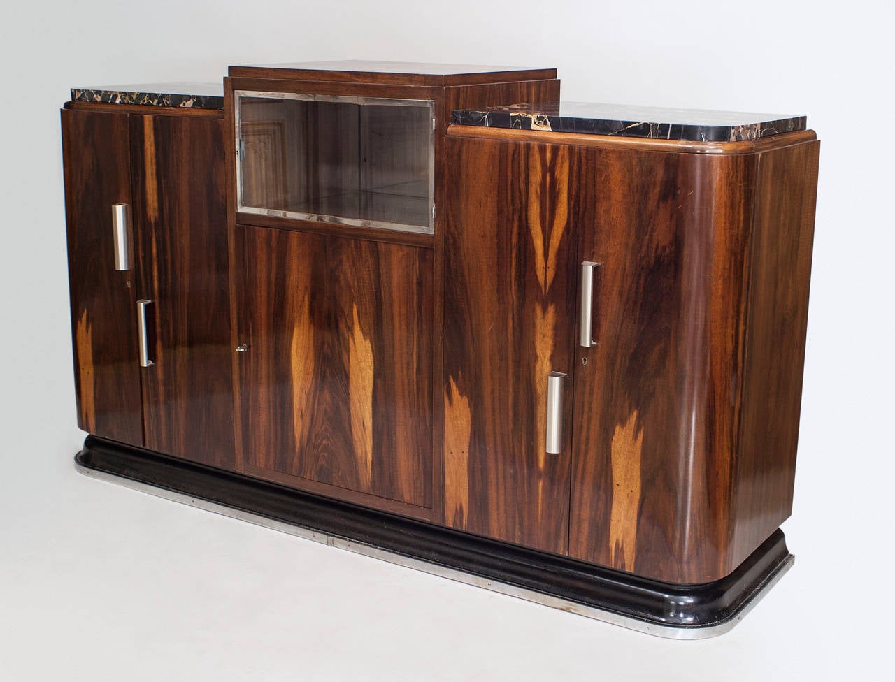 French Art Deco rosewood sideboard with a raised center platform over a glass door shelf centering 2 black marble tops. Doors with chrome handles and trim.
