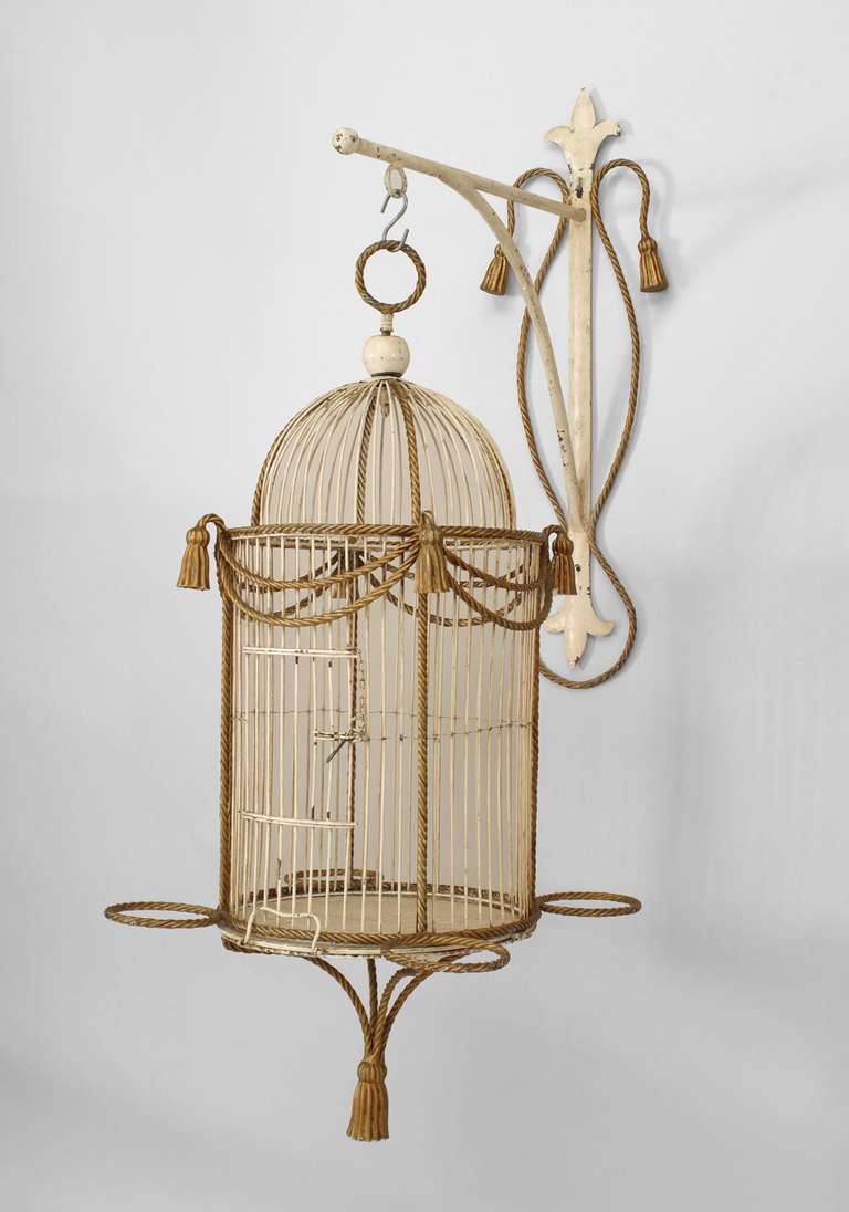 The cylindrical Italian birdcage composed of white and gold painted metal in a decorative rope and tassel style. The birdcage can be suspended from a crowning ring as well as an optional wall bracket.
