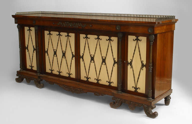 English Regency rosewood sideboard featuring a brass gallery, filigree back rail, scroll-form legs, and four doors faced with cream upholstery and diamond-shaped bronze grills.