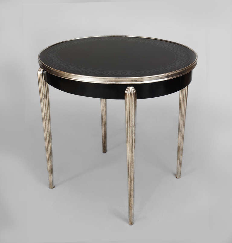 French Art Deco style end table with an inset black oval glass top with a silver gilt rim and reverse silver painted border resting upon silver gilt fluted legs and upper rim.