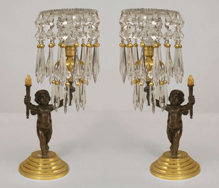 Pair of French Charles X left and right candlesticks cast in the form of bronze cherubs standing upon tiered round gilt bases and elevating small torches with one arm and crystal and gilt candleholders in the other.