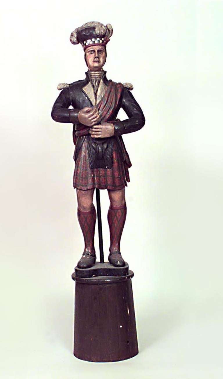 Large nineteenth century large carved and painted wooden figure representing a Scottish Highlander wearing a traditional plumed cap and uniform with kilt, blazer, and tartan sash. The figure is propped upon a large round base.