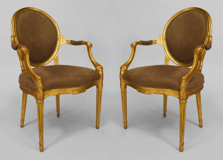 Pair of English George III arm chairs dating to 1780 and crafted in the French Louis XVI style. Each armchair features a frame composed entirely of giltwood with an oval back and out turned arms over four tapered fluted legs. The seats, backs, and