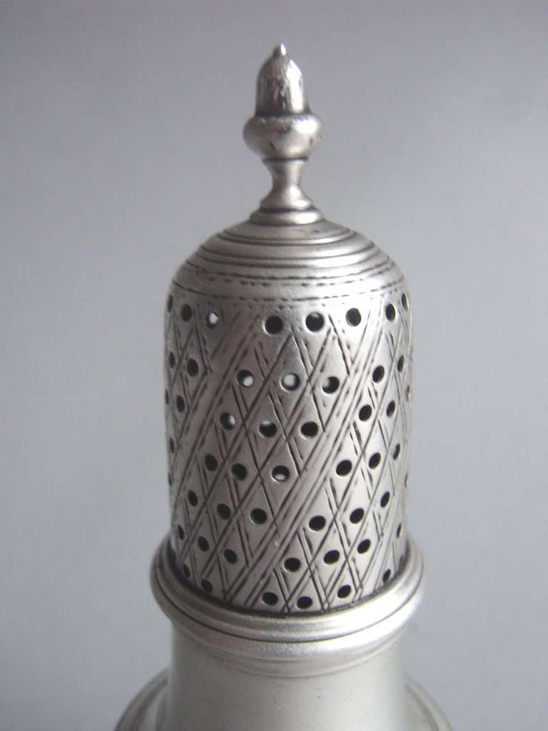 The Caster stands on a circular pedestal foot which is decorated with reeding, as is the baluster shaped main body. The pull-off cover is pierced with roundels and is engraved with trellis work and terminates in an acorn finial. The Caster is fully