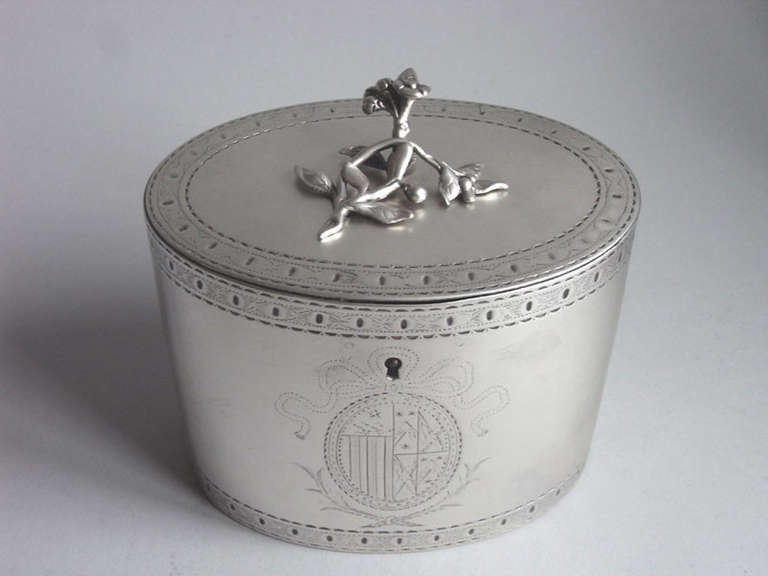 The tea caddy is oval in form and the sides of the main body and edge of the cover, are engraved with bright-cut and scratch engraved bands. The front of the caddy displays a contemporary Armorial surrounded by a bright cut cartouche with tied