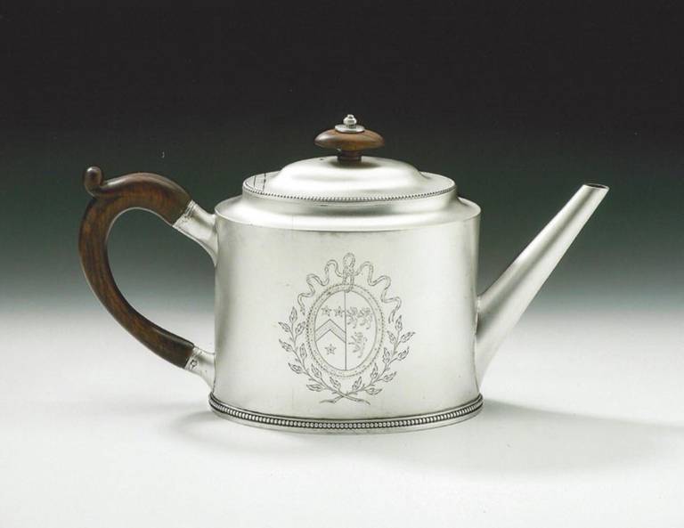 The teapot is modeled in the straight sided oval form with a beaded band at the base. The incuse top has a beaded rim and rises to a slightly domed hinged cover which terminates in a silver and fruitwood finial. This piece has a straight spout and