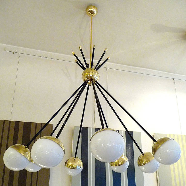 A beautiful chandelier featuring a brass sphere with eight blackened arms, all of which diagonally penetrating out the middle sphere. Each arm terminates in a brass cup which holds a white opaque glass ball shaped light diffusing shade.