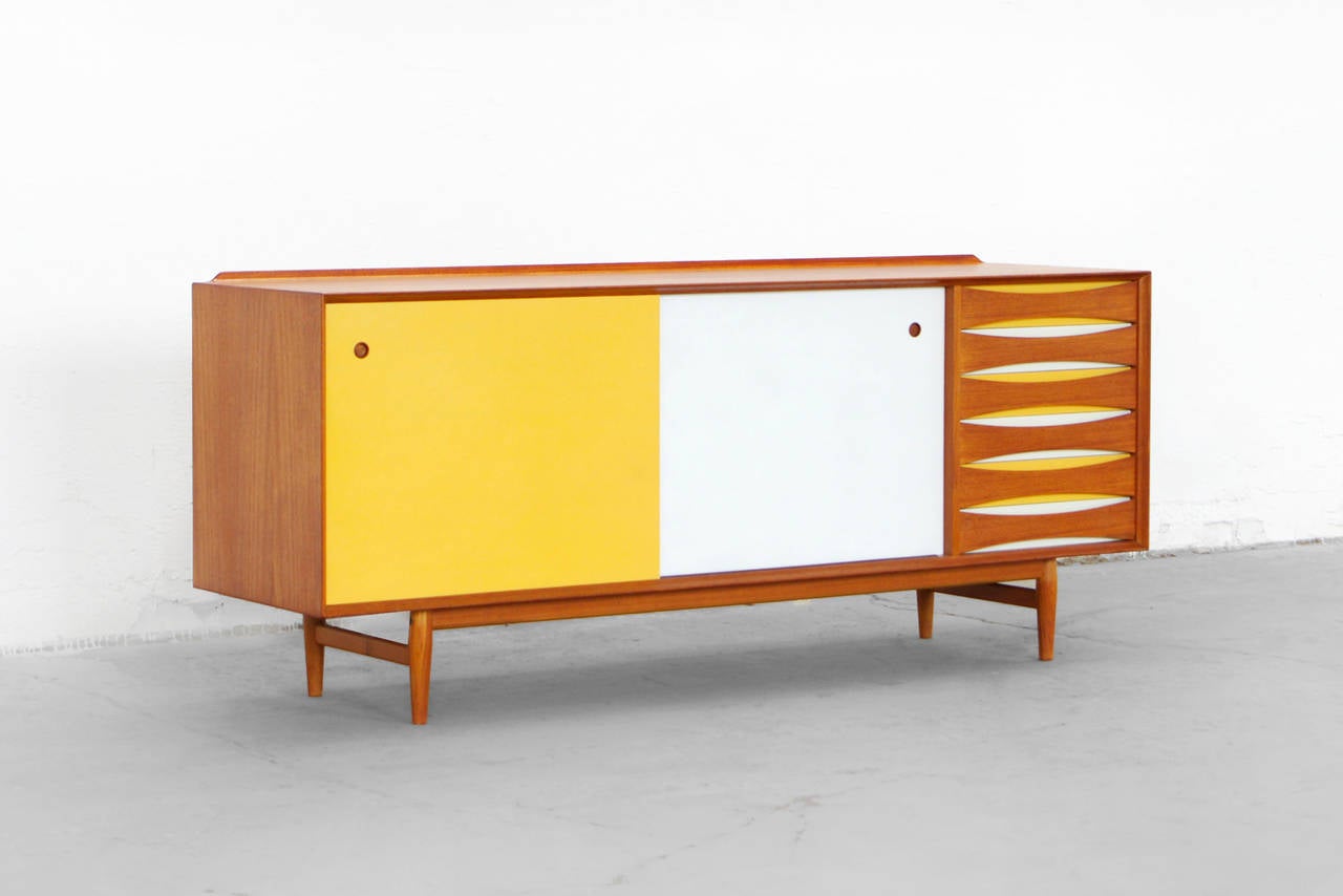 Arne Vodder's timeless OS 29 freestanding sideboard with colored cabinet fronts. The sliding doors are reversible - with teak on the one side and white and yellow lacquer on the other. 
The piece offers great storage options with drawers and