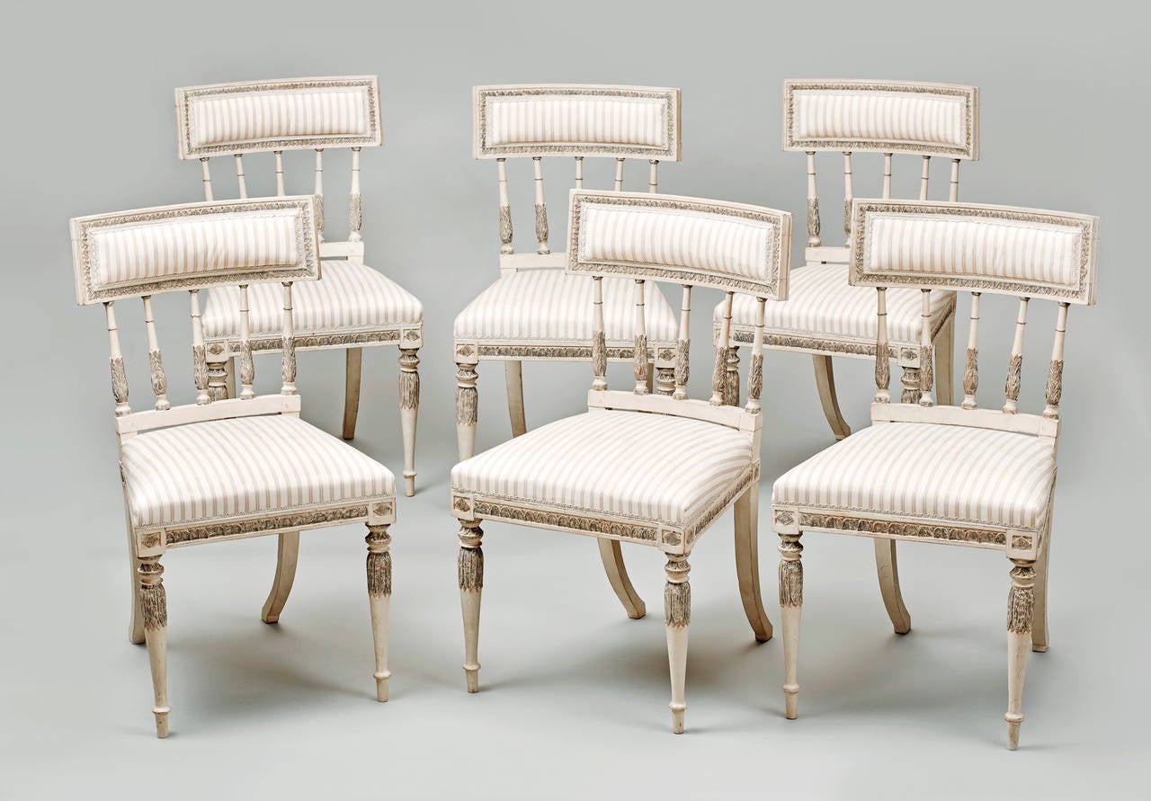 An elegant set of six Swedish Gustavian dining chairs from the neoclassical period painted in cream white and grey, with leaf tip carvings overall. The front round tapering legs with leaf carving, to the back sabre legs. Upholstered seat and back