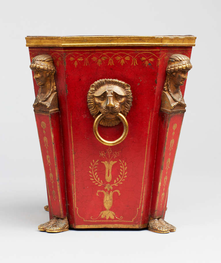 An Empire tole peinte ormolu-mounted jardiniere or planter from the early 19th century. Painted with gilt empire ornaments to all sides against a red ground, each corners with a classical caryatide in gilt-bronze, with two lion mask side handles,