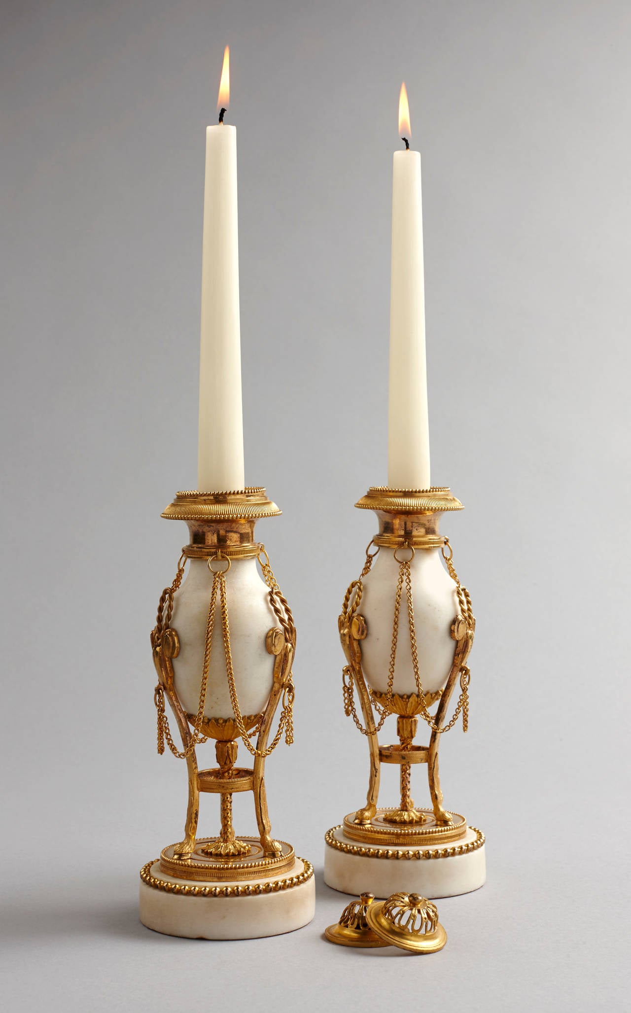 A fine pair of Louis XVI white marble and gilt bronze cassolettes in the form of atheniennes. Each with a domed and pierced cover to reveal the candleholder, the egg ovoid shaped white marble body with ormolu chain swags and supported by three hoove