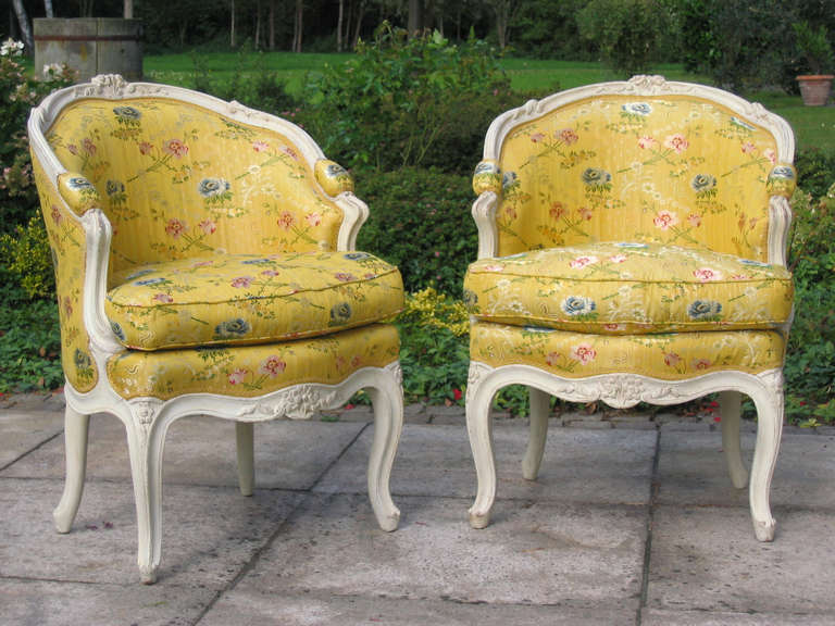 A pair of Louis XV white painted Bergeres en corbeille, circa 1755. Each with padded back and seat with channelled frame and foliate-carved rails and cresting, with padded arms, on cabriole legs, upholstered in yellow silk (‘Lampas Poussin’ by