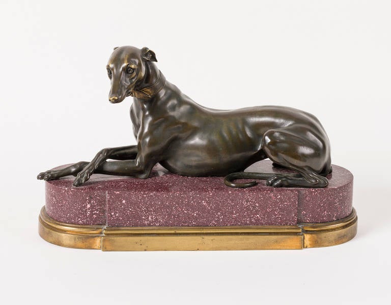 A decorative bronze figure of a greyhound, French, first half of the 19th century. Seated, her forelegs crossed, her head modelled with attentive expression, dark brown patina, on a shaped porphyry base with gilt bronze plinth.