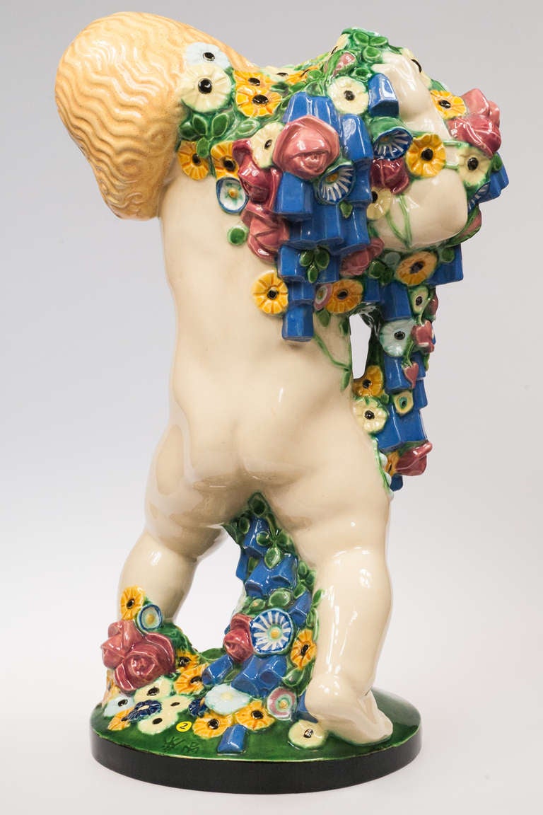 This item is a pottery putto designed by Michael Powolny. It represents one of the four seasons, the spring. The putto is holding a bunch of flowers, which have a magnificent variety of colors. There are two marks at the bottom and in the green, the