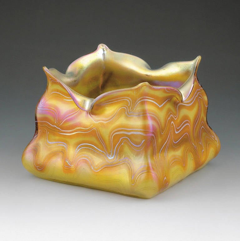 Bowl, 1902
Colourless glass, overlayered with yellow, wavy combed silver-yellow threaded and red banded décor, with strong matt gold and pearl textures, square bottom, off set wall and strongly articulated quatrefoil mouth
The polished cut-off
