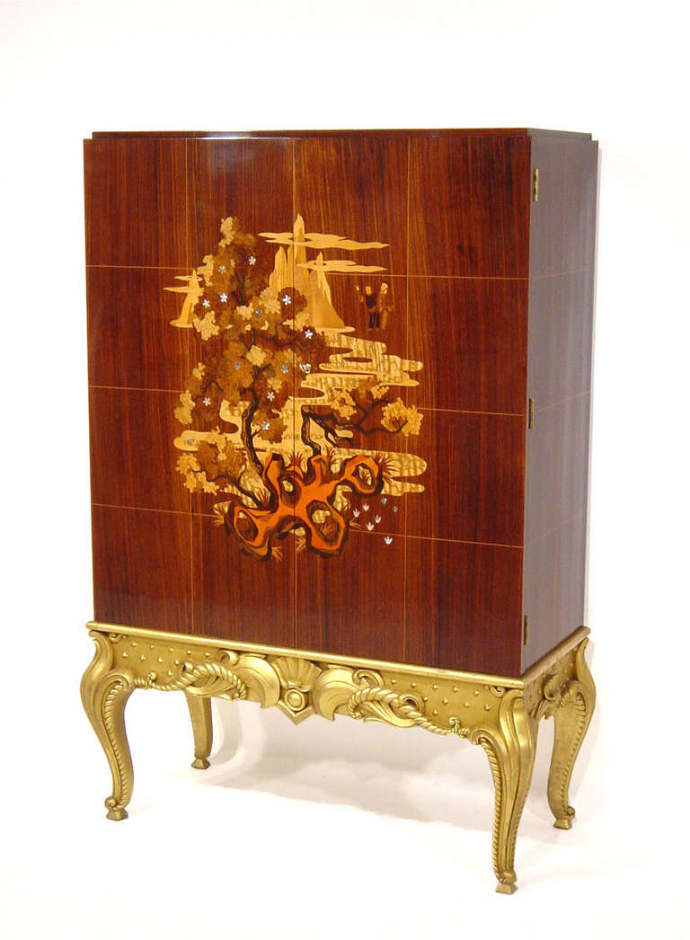 A fine cabinet with a gilded base and and a rosewood corpus. In the center this marvelous item is decorated with inlays of very high quality. Different kinds of precious woods and mother-of-pearl form a chinese-inspired scene with a beautiful
