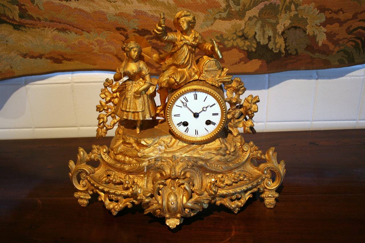 This Napoleon III regule doré mantel clock features a courting couple. The boy is raising a glass of champagne in one hand, and holds the bottle in his other, while the young lady holds her parasol. He is leaning on a wine barrel, which holds the