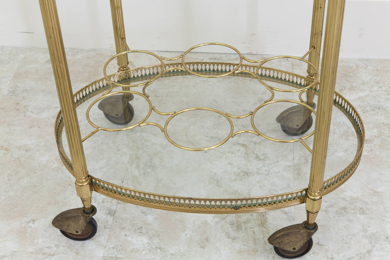 This mid-20th century polished brass bar cart with pierced gallery features a handled removable glass tray for serving and a lower shelf with bottle holders for storage of six bottles. The wheels roll smoothly, making this piece an elegant and